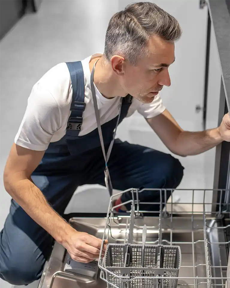Reparatur oder Neukauf Bild zeigt Handwerker, der Spülmaschine öffnet, während er davor hockt.